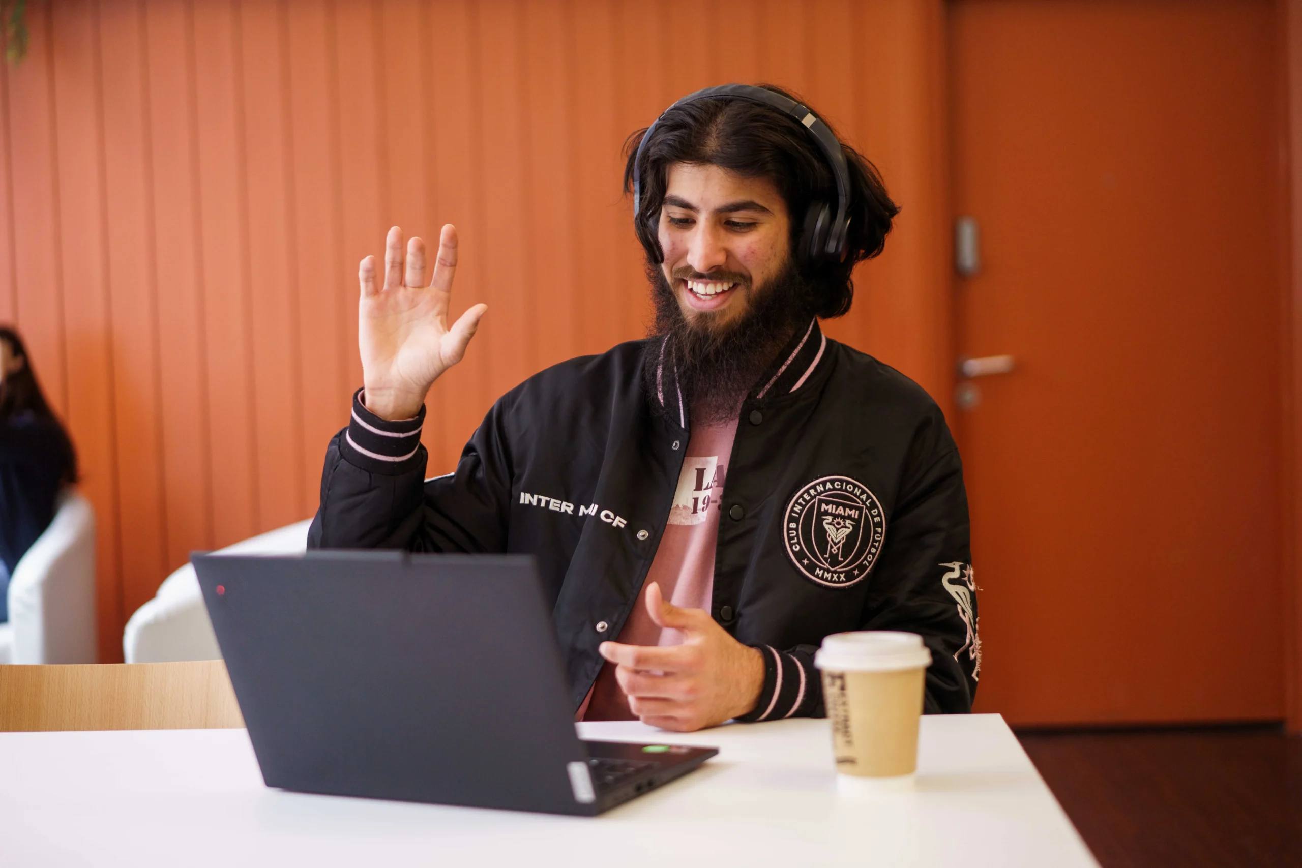 How online education prepares students for emerging industries - bearded man with headphones on and takeaway coffee raising hand in a call on laptop