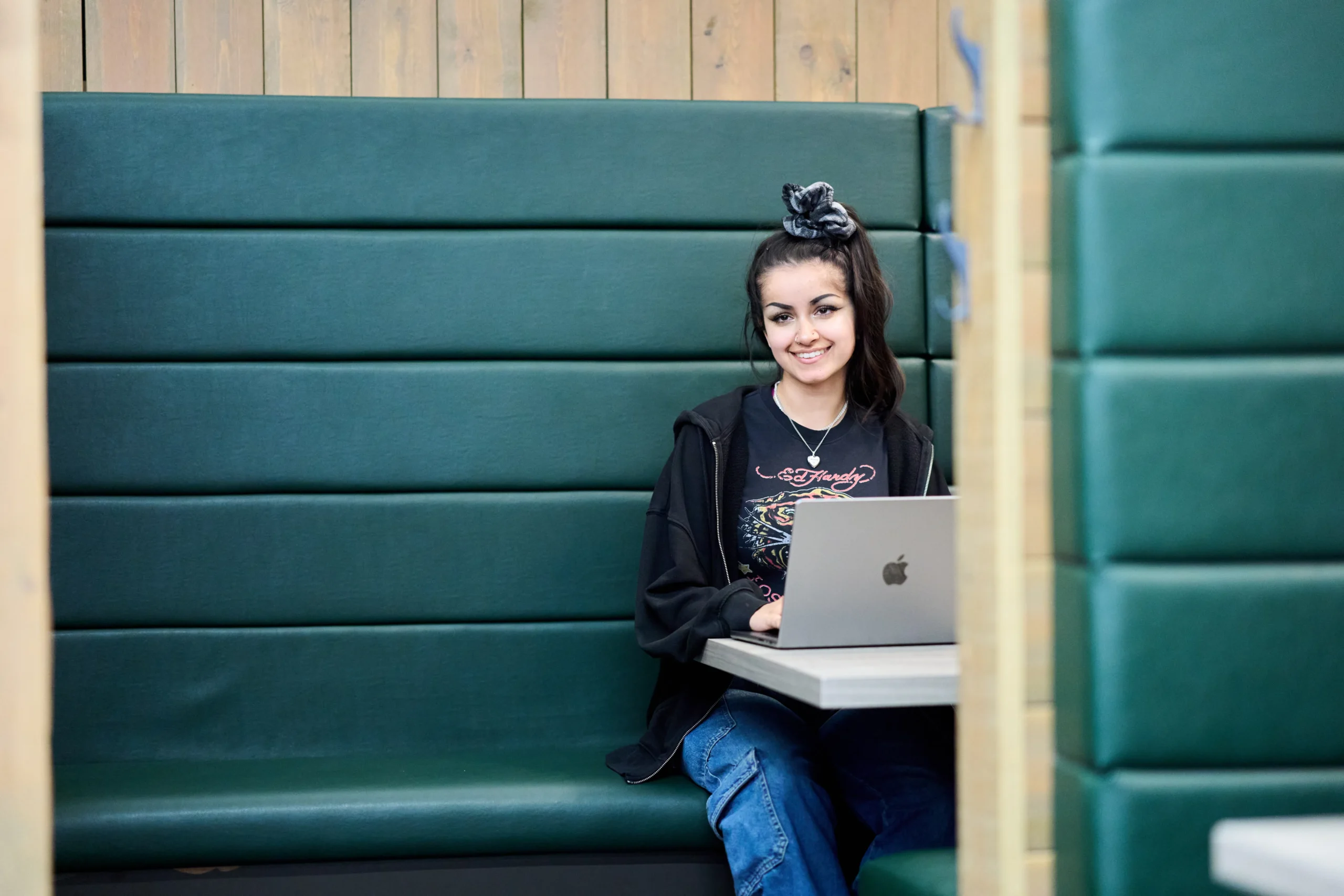 How DBA and PhD students in business differ - woman sitting alone in a green booth in front of mac computer smiling at camera