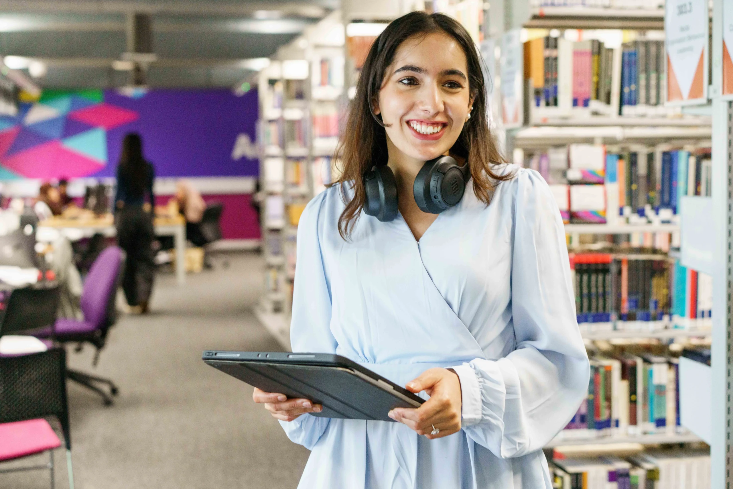 How to become a lawyer with an Aston Online Bachelor of Law LLB (Hons) Degree - smiling woman in library holding tablet with headphones around neck