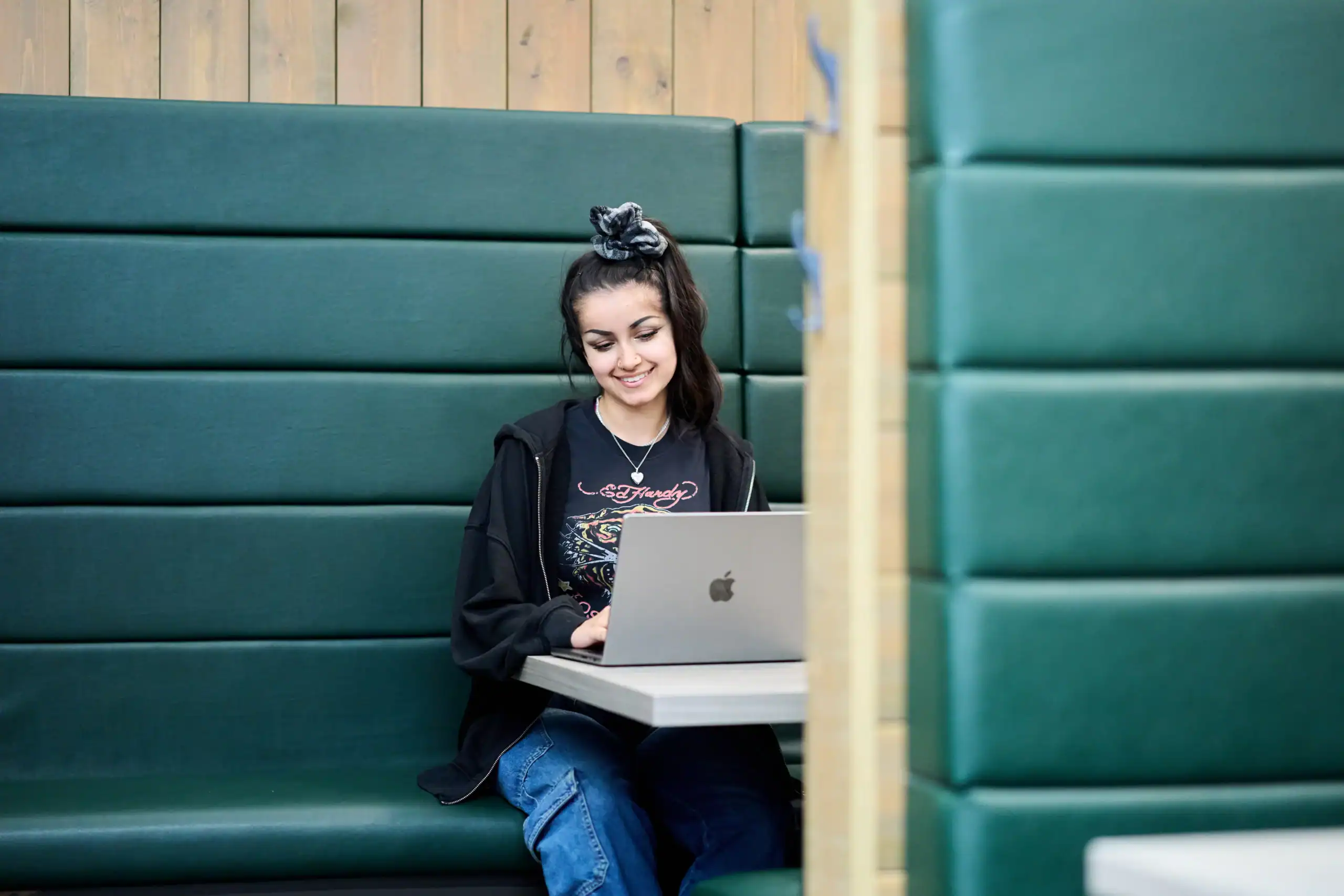 Select your Aston Online Programme - woman sitting in booth smiles while looking down at laptop